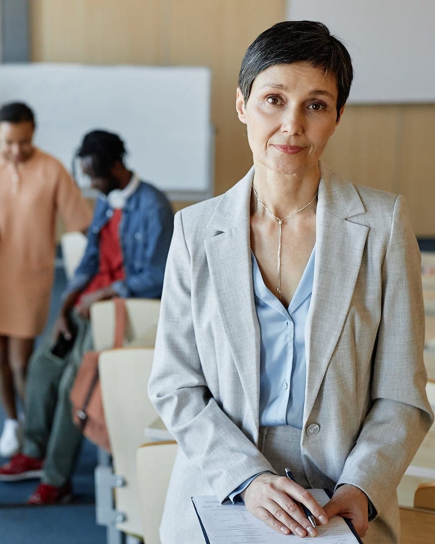 A senior leader in a light grey suit stands in a lecture theatre, students talking together behind her