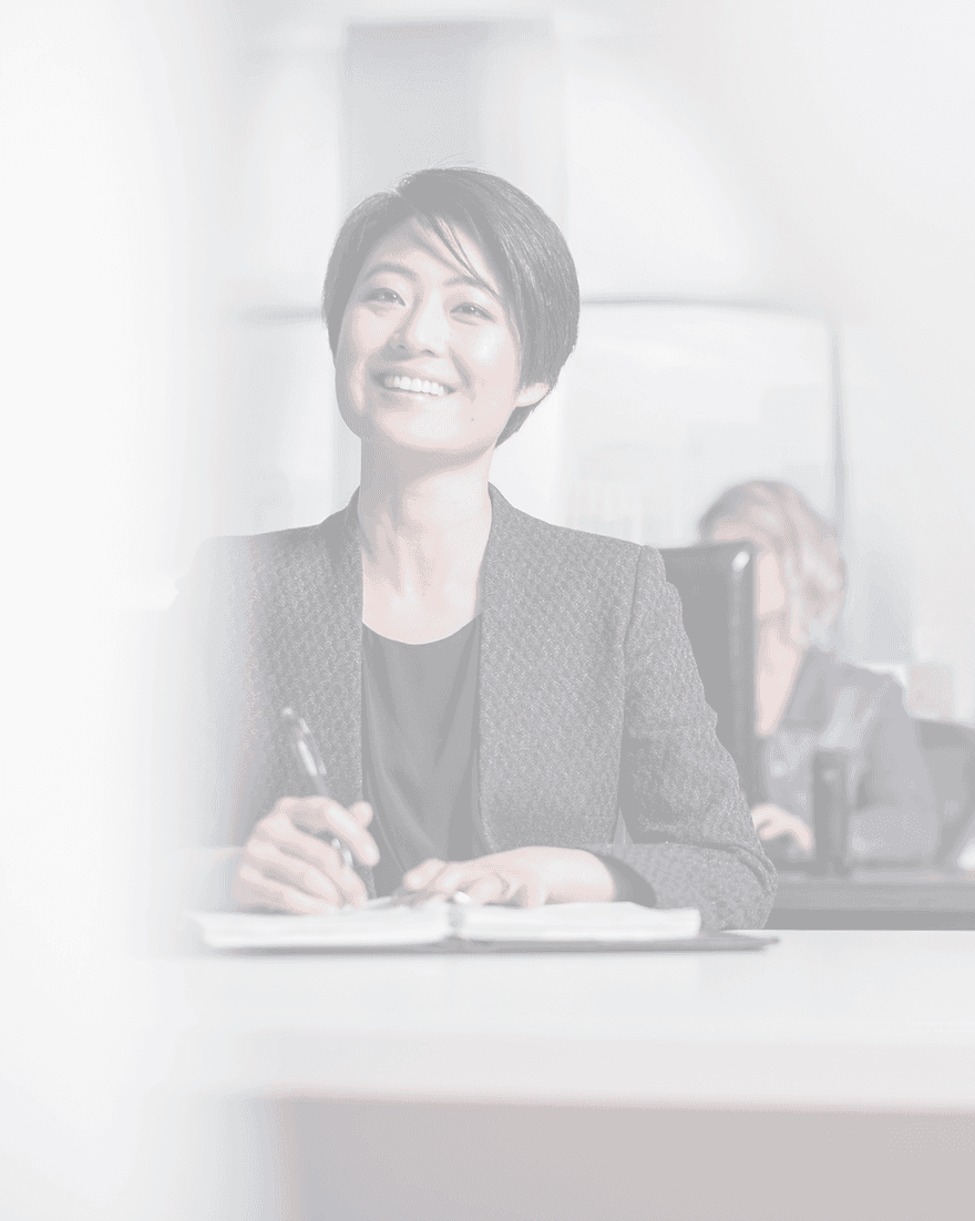 A smiling executive in a dark blazer sits at a boardroom table making notes