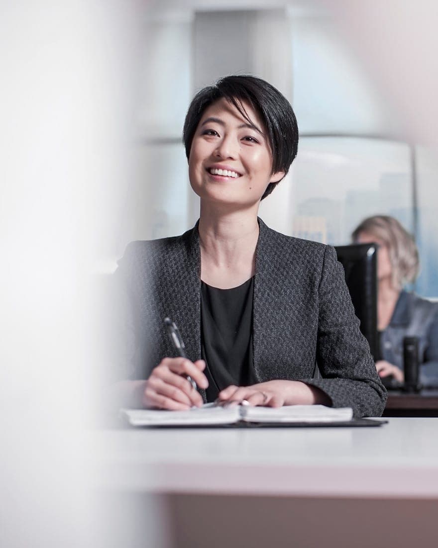 A smiling executive in a dark blazer sits at a boardroom table making notes